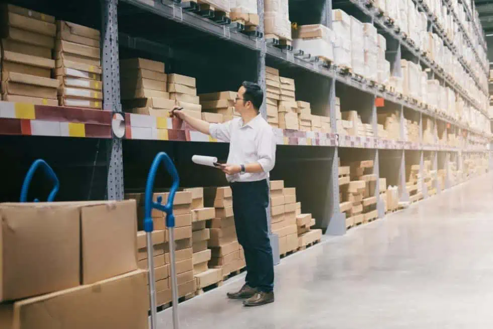 In a bustling warehouse, a person in a white shirt jots down notes on a clipboard. They are surrounded by shelves packed with boxes, representing the diverse sectors eRep Industries proudly serves. A hand truck stacked with even more boxes waits in the foreground.