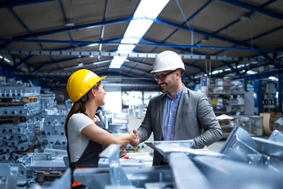 Two people wearing hard hats shake hands in a bustling factory setting surrounded by metal structures, embodying the collaborative spirit of eRep Industries we serve.