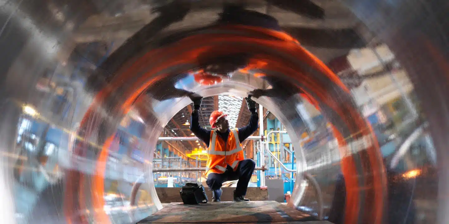 A worker in a hard hat and safety vest inspects the interior of a large reflective metal pipe on a construction site, ensuring everything is ready for the CPQ pump selector system installation.