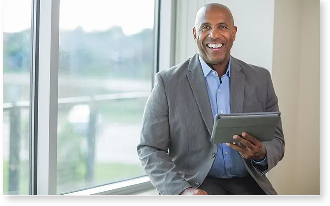 A smiling man in a suit sits by a window, holding a tablet with revalize support, as the blurred outdoor scenery adds to his focused demeanor.