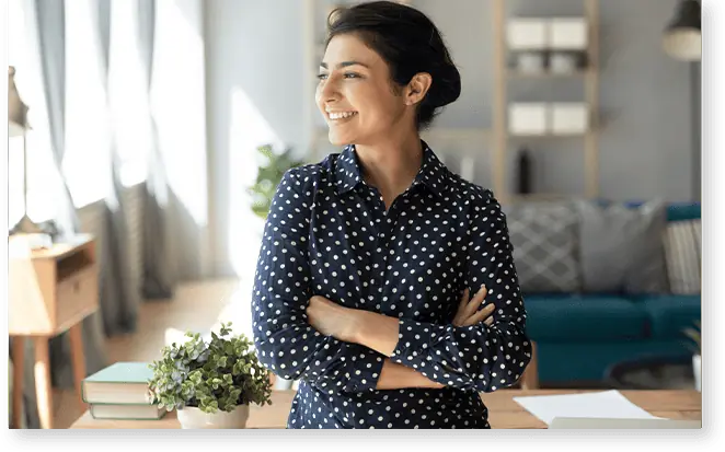 Woman in a polka dot shirt smiles confidently, standing with arms crossed in a bright room with bookshelves, exuding the kind of warmth and revalize support that makes everyone feel at ease.