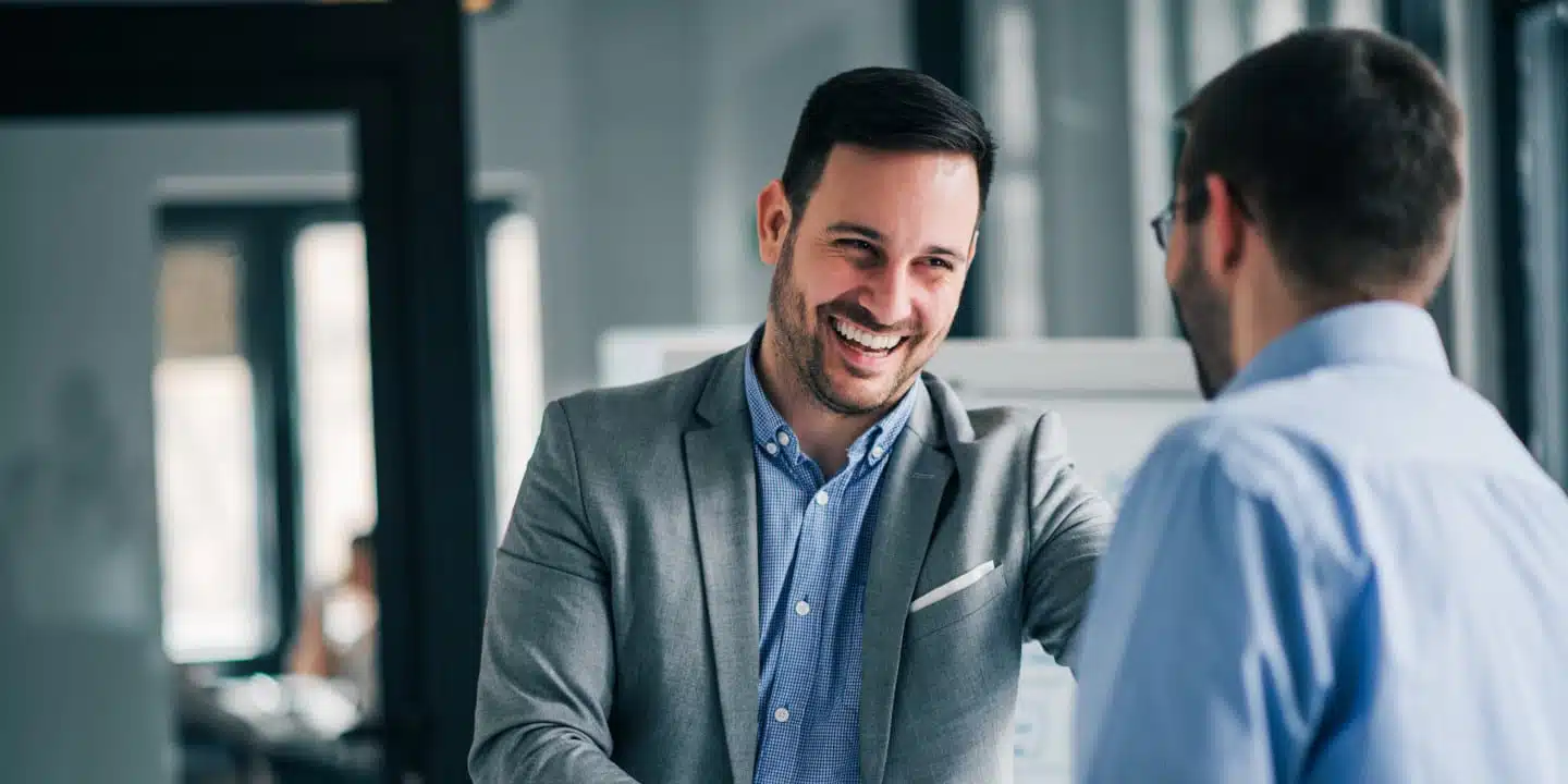 Two men are having a conversation in an office about choosing the right CPQ. One man, smiling in a gray suit, listens intently while the other, wearing a blue shirt, gestures animatedly.