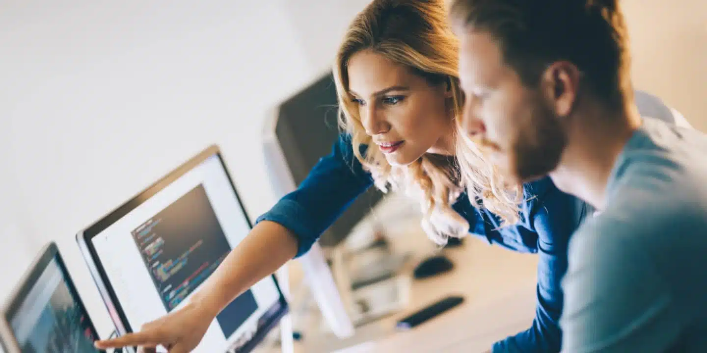 Two people working at a desk with multiple computer monitors, one pointing at the screen displaying code related to CPQE and PLM solutions.