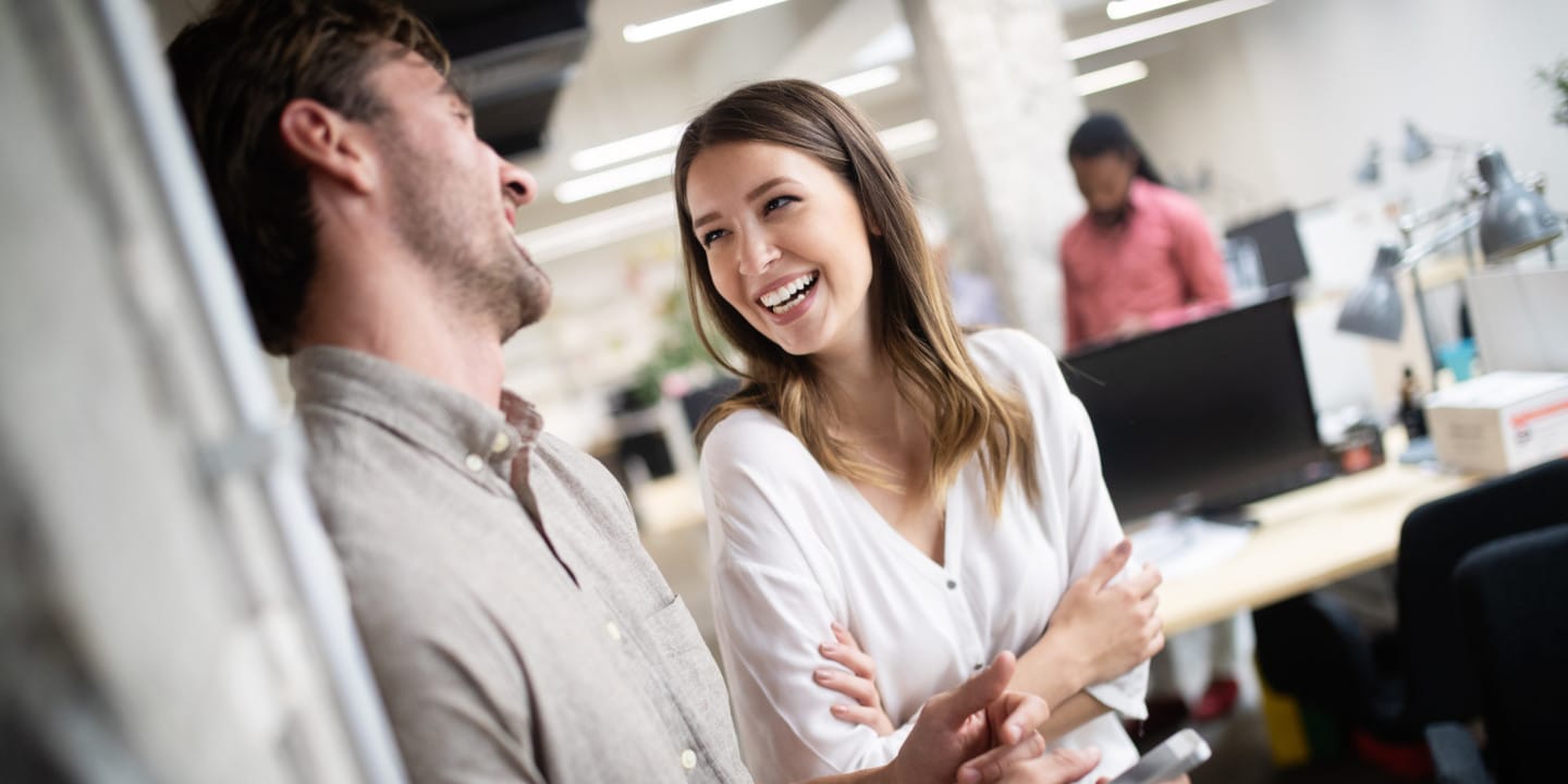 Two people, debunking CPQ misconceptions, laugh and chat in a bright, modern office. One leans against a wall while the other stands with arms crossed. Desks and computers are scattered in the background, creating an atmosphere of innovation and collaboration.