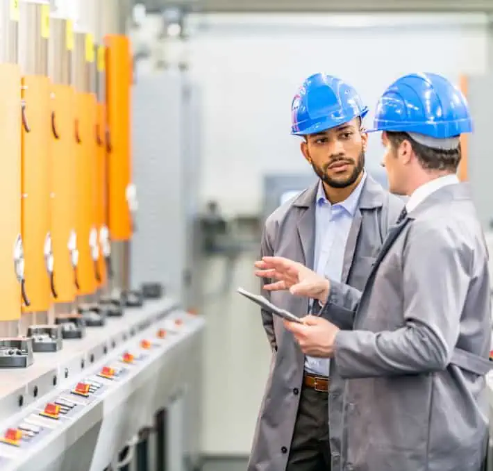 Two men wearing blue hard hats and gray jackets discuss in a factory setting, surrounded by industrial equipment with orange panels. One holds a tablet, likely using the Intelliquip CPQ system to streamline their workflow.