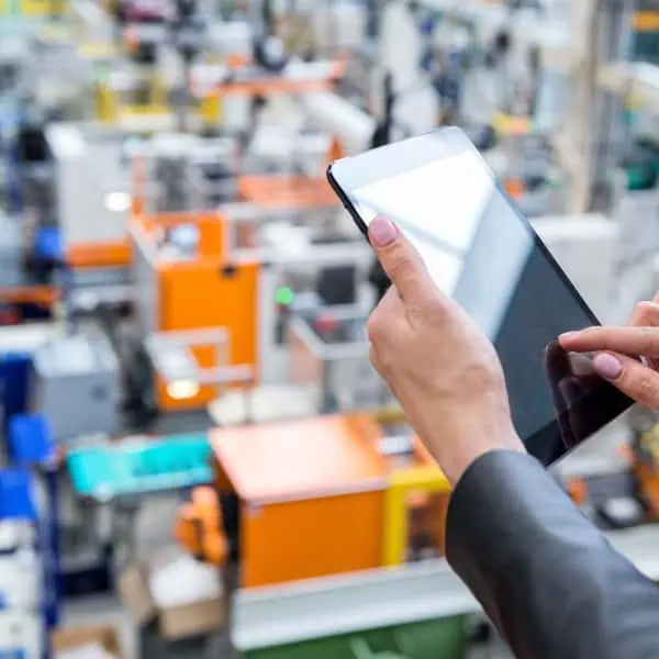 Amidst the hum of machinery, a person holds a tablet in a factory setting, seamlessly integrating Intelliquip CPQ to enhance workflow efficiency.
