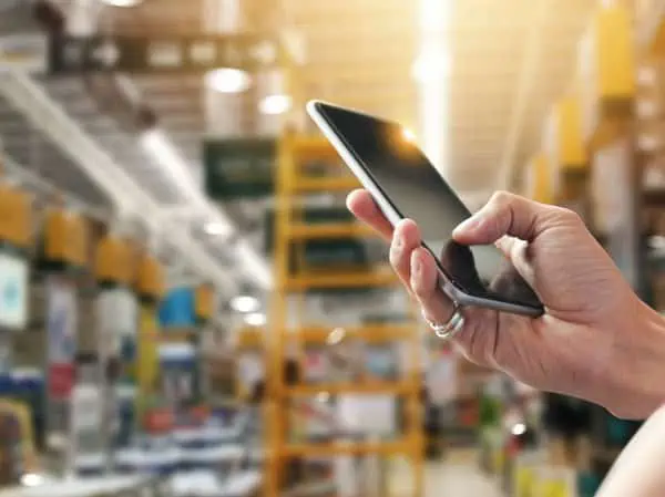 In a brightly lit store aisle, a person expertly navigates the Intelliquip Project and Quote Management Portal on their smartphone. Shelves and products blur into the background, emphasizing their focus on efficiently managing tasks at hand.