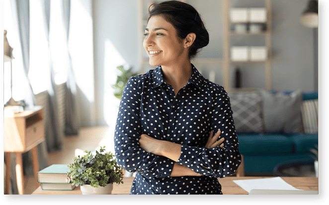 Woman in a polka dot shirt smiles confidently, standing with arms crossed in a bright room with bookshelves, exuding the kind of warmth and revalize support that makes everyone feel at ease.
