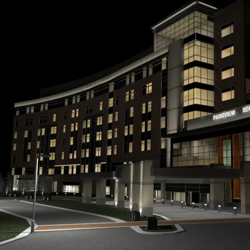 A modern multi-story building with many windows is illuminated at night, with exterior lights—designed using Illuminating Engineering Software—highlighting the entrance and surrounding pathways.