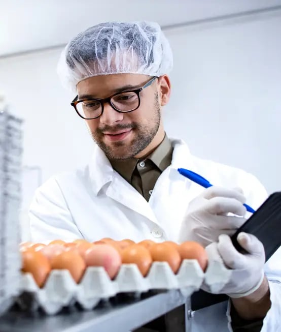 A person in a lab coat and hair net examines eggs, holding a clipboard and pen.