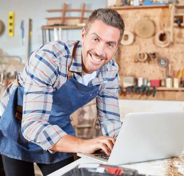 Man in a workshop wearing a blue apron, using a laptop on a workbench.