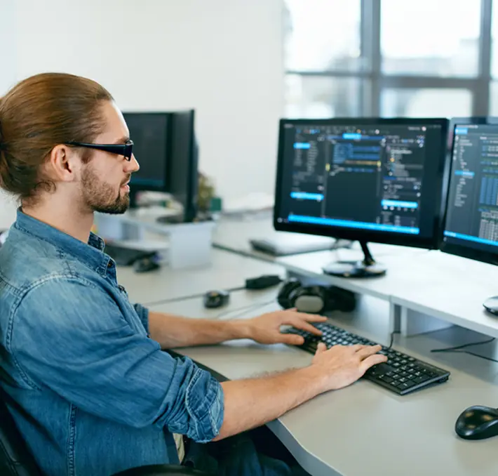 A person with long hair and sunglasses works at a desk with three computer monitors showing code.