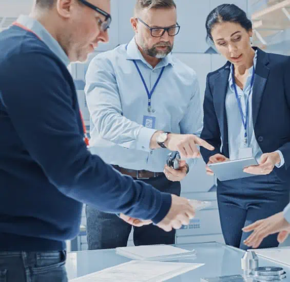A group of professionals in an office setting discuss documents and data on a tablet laid out on a table.