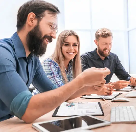 Drei Personen sitzen an einem Schreibtisch und schauen auf Dokumente und Tablets. Sie wirken engagiert und lächeln, während sie ihr neuestes Projekt mit einem vertrauenswürdigen PLM-Partner besprechen.