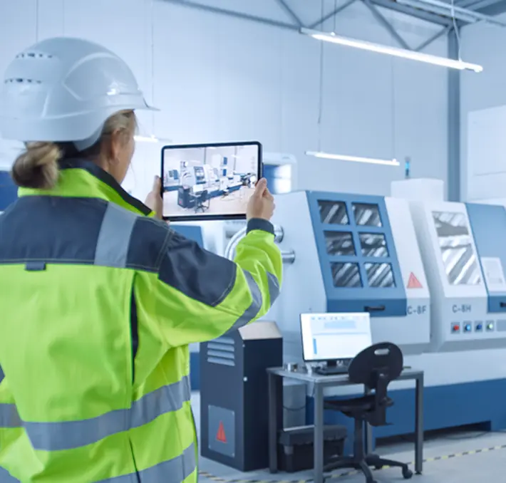 A person in a high-visibility jacket and helmet uses a tablet to capture images in a factory with machinery and monitors in the background.