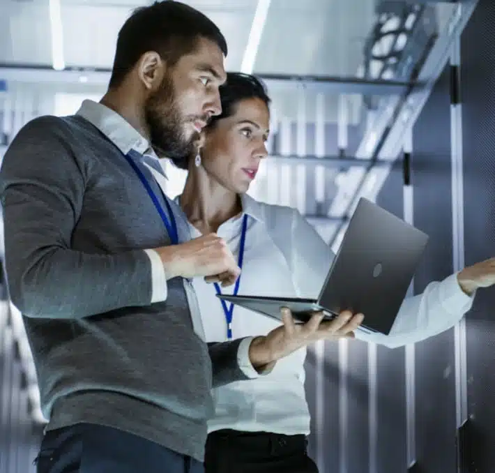 Two people in a server room examining equipment as part of engineering change management. One holds a laptop, and both wear ID lanyards, focused on ensuring updates are seamlessly integrated.
