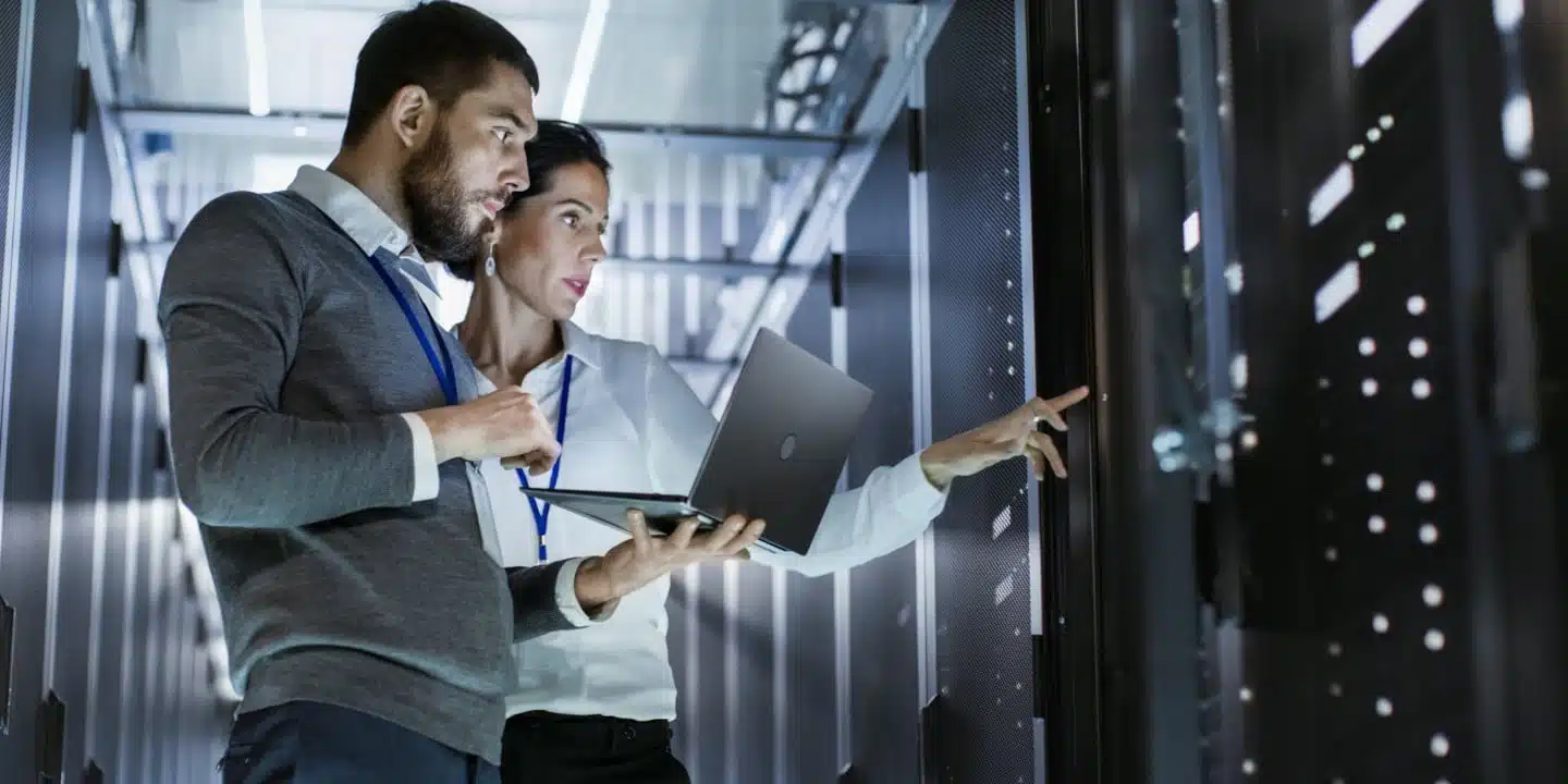 Two people examine server racks in a data center, one holding a laptop while the other points at a server. Theyre engaged in engineering change management, ensuring efficient updates. Both wear identification badges.