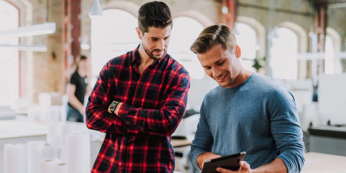 Two men in casual clothing are standing indoors. One in a red plaid shirt is observing, while the other in a blue sweater, perhaps a PLM partner, is using a tablet. They appear to be in a modern workspace.