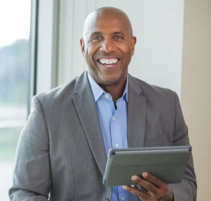 Smiling man in a suit holding a tablet, sitting near a large window.
