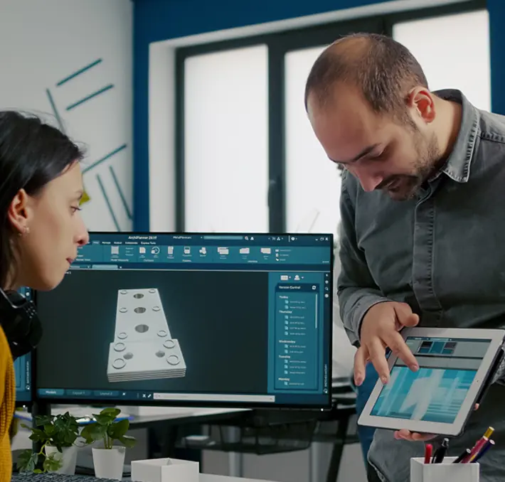 Two people in an office discuss 3D model designs displayed on a computer and tablet. A monitor shows a 3D object.