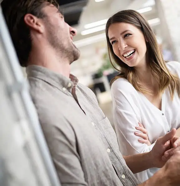 A man and a woman are standing together, smiling and laughing in a brightly lit indoor setting, embodying the cheerful spirit of Scranton Product.