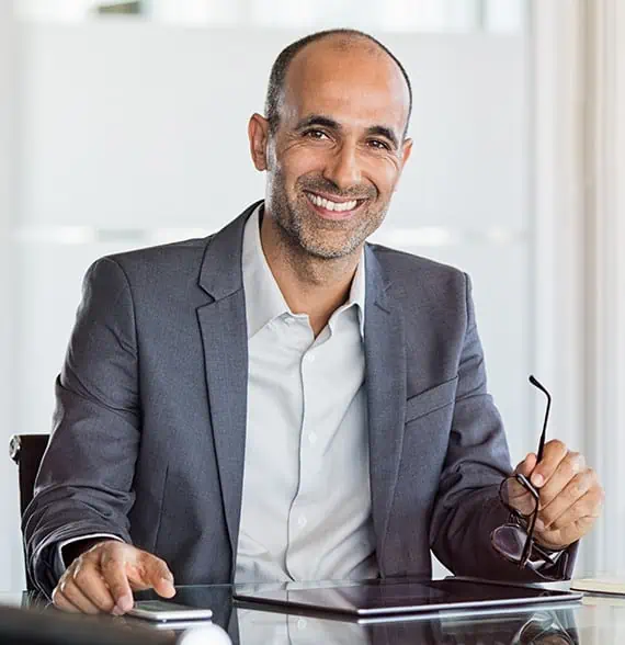 A man in a gray suit sits at a desk, smiling and holding glasses, much like the warm welcome youd receive at Tommy Car Wash.