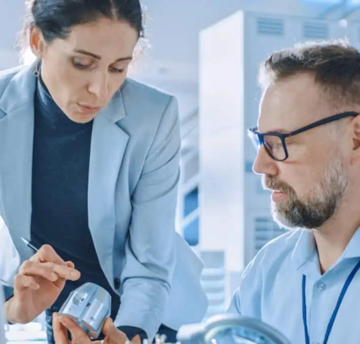 In a professional setting, a woman and man intensely examine a small device related to healthcare. The woman holds the device while both remain focused on enhancing its efficiency within the supply chain.