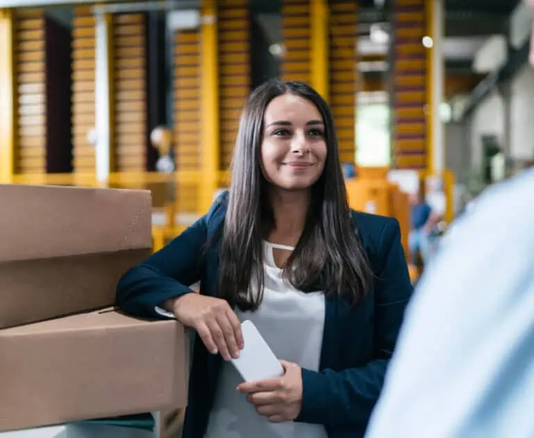 In a bustling warehouse setting, a woman in business attire leans on a stack of boxes emblazoned with the Dr. Oetker logo, expertly balancing her smartphone as she orchestrates logistics with impressive efficiency.