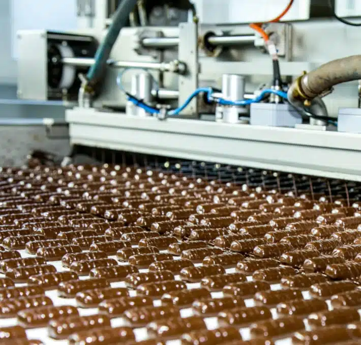 In einem Schaufenster der Lebensmittelinnovation befördert ein Fließband zahlreiche Schokoladenbonbons, die in einer geschäftigen Fabrikumgebung von hochmodernen Maschinen fachmännisch verarbeitet werden.