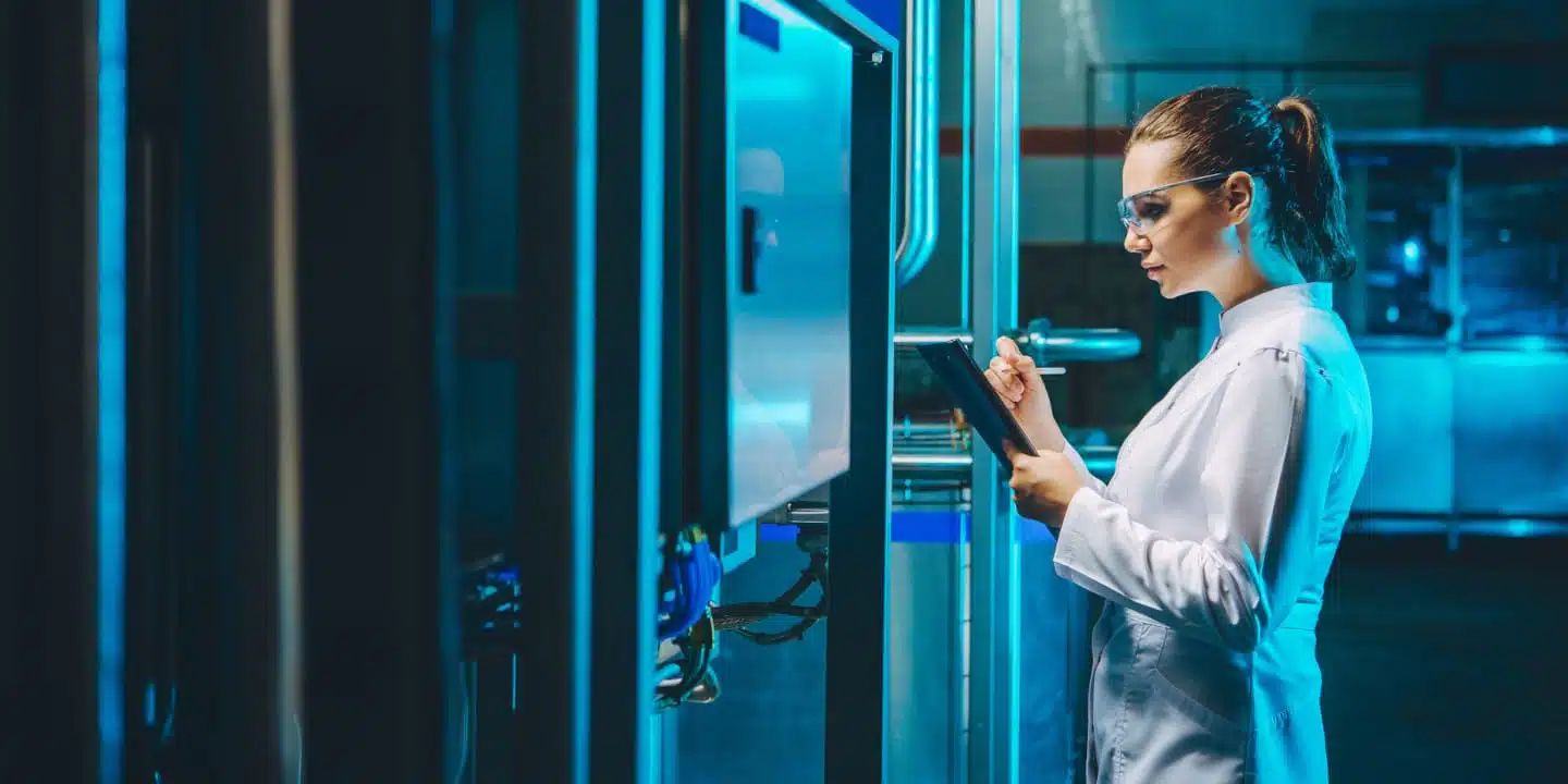 A person in a lab coat and safety glasses checks a control panel, ensuring precision in the food & beverage industry, and takes notes on a clipboard in an industrial setting.