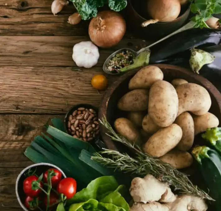 A wooden table showcasing a vibrant assortment of vegetables: potatoes in a bowl, tomatoes, ginger, garlic, rosemary, onions, eggplant, and green leaves alongside a spoonful of mixed spices—reflecting the spirit of PLM for food & beverage innovation.