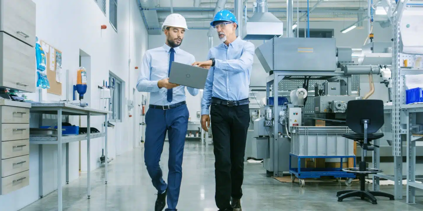 Two men in hard hats and shirts walk through an industrial facility discussing a laptop. One wears a white hard hat, the other a blue one. Machinery is visible in the background.