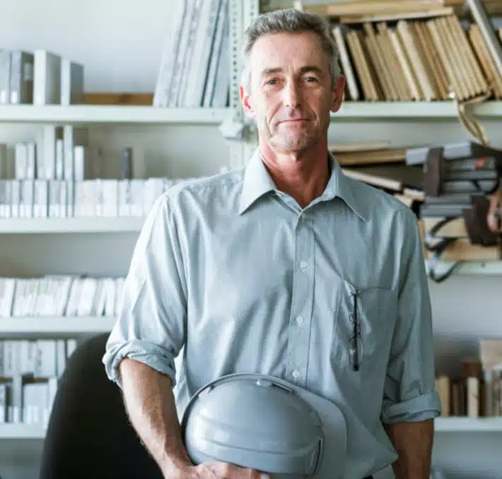 A man in a gray shirt holds a gray hard hat, standing in an office with shelves filled with an extensive medical equipment catalog alongside books and files.
