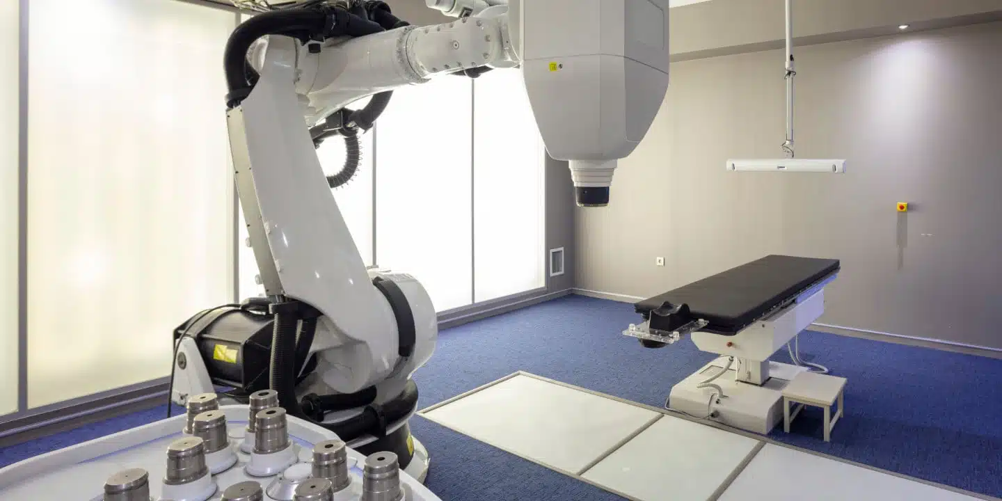 A robotic medical device, part of the streamlined medical equipment TCO strategy, is positioned near an examination bed in a modern, well-lit room with blue carpeting and beige walls.