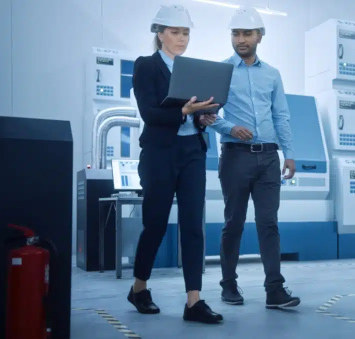 Two people wearing hard hats walk through a modern industrial facility. One holds a laptop, while machinery, including medical equipment and a fire extinguisher, are visible in the background.