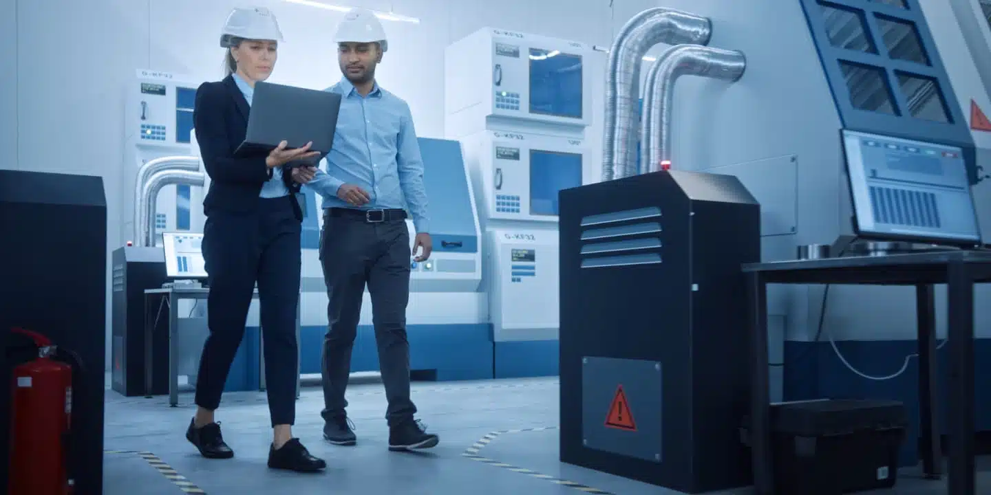 Two engineers in hard hats review data on a laptop, considering the total cost of ownership (TCO) for medical equipment, as they walk through a modern industrial facility.