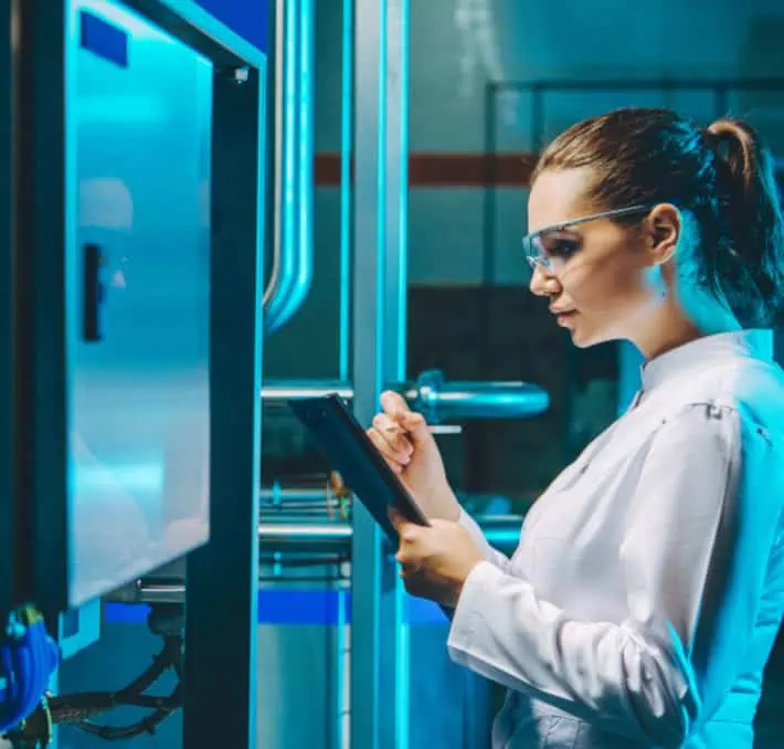 A woman in a lab coat and safety glasses uses a tablet while standing next to industrial equipment with blue lighting.