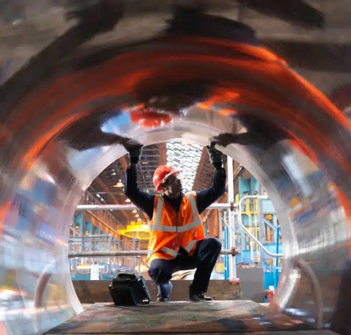 A worker in an orange vest and hard hat inspects the interior of a large metallic pipe in an industrial setting, ensuring compatibility with CPQ pump selector standards.