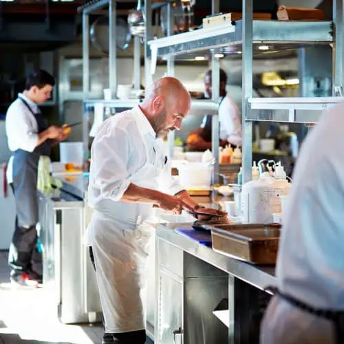 Chefs in a professional kitchen prepare dishes with precision. At a stainless steel counter, one chef meticulously plates food, utilizing insights from food and beverage manufacturing software, while others skillfully work in the background.