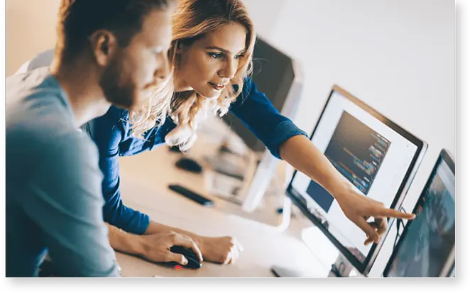 Zwei Personen arbeiten an einem Schreibtisch, konzentriert auf ihre Bildschirme. Der eine zeigt auf den Monitor, bietet dem anderen seine Unterstützung an und schlägt ihm eine Idee vor, um die Zusammenarbeit zu fördern.