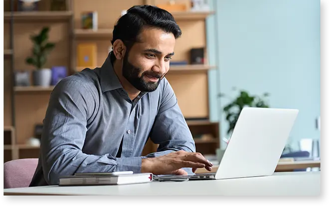 Ein Mann in einem grauen Hemd tippt an seinem Schreibtisch auf einem Laptop und verkörpert so perfekt den Revalize Support, während er von Büchern und einer Pflanze umgeben ist, die den Hintergrund charmant ergänzen.