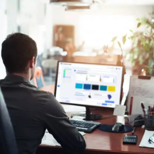 Eine Person sitzt an einem Schreibtisch, den Blick auf einen Computermonitor gerichtet, auf dem eine diskrete Fertigungssoftware angezeigt wird. In der Nähe befinden sich Büromaterialien und eine Pflanze, die einen ausgewogenen und effizienten Arbeitsbereich schaffen.