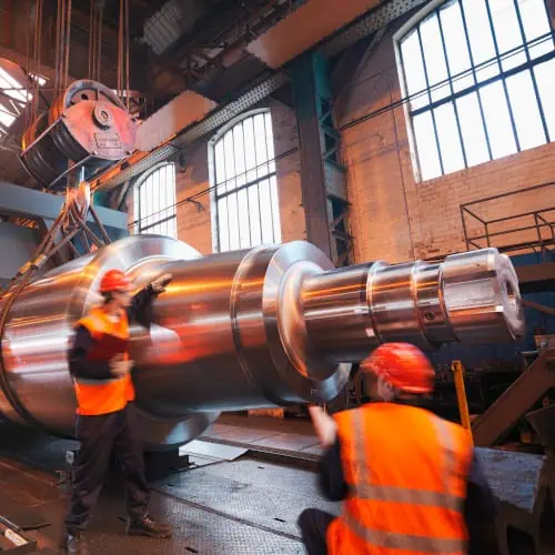 Zwei Arbeiter in orangefarbenen Sicherheitswesten und Helmen inspizieren einen großen, glänzenden Metallzylinder in einer industriellen Umgebung mit großen Fenstern. Sie verwenden eine diskrete Fertigungssoftware, um Präzision und Effizienz zu gewährleisten.