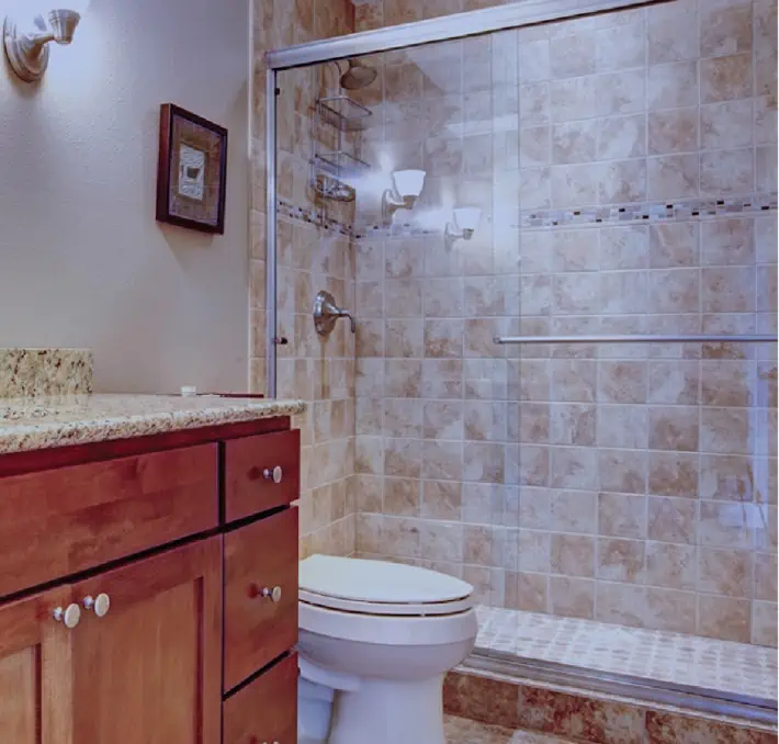 Modernes Badezimmer mit Holzschränken, einer Arbeitsplatte aus Granit und einer Dusche mit Glasabdeckung von Quality Enclosures und beigen Fliesen. Die weiße Toilette befindet sich vor der Dusche. Darüber befindet sich eine wandmontierte Leuchte.
