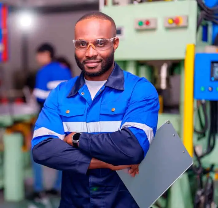 A man in a blue work uniform and safety glasses stands with crossed arms, holding a clipboard in a industrial setting.