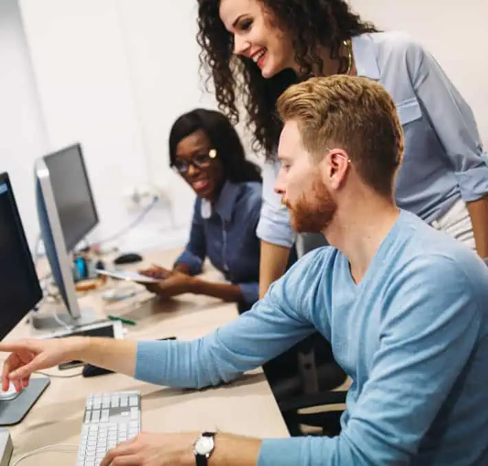 Three people collaborate at a desk with computers, using Salesforce tools. A man points at the screen, while one woman stands and another sits, all engaged in vibrant discussion.