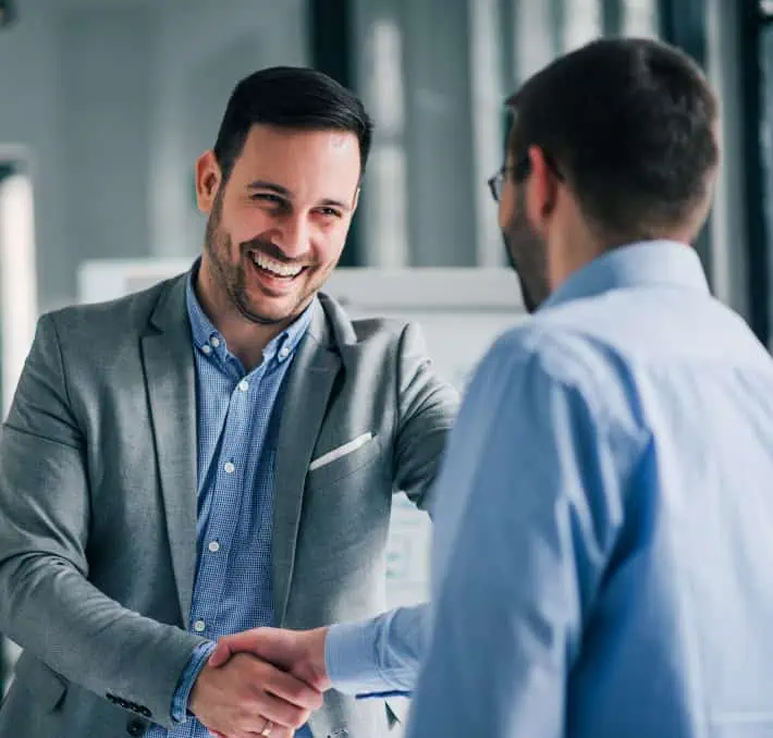 In an office setting, two men in business attire shake hands and smile, confidently deciding on choosing the right CPQ to elevate their business solutions.