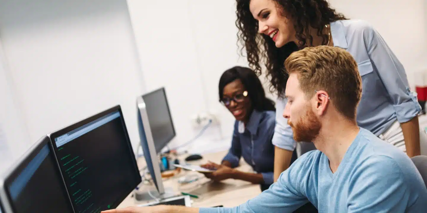 Three people collaborating at a computer workstation, with one person typing on a keyboard while the others observe and discuss strategies using Salesforce.