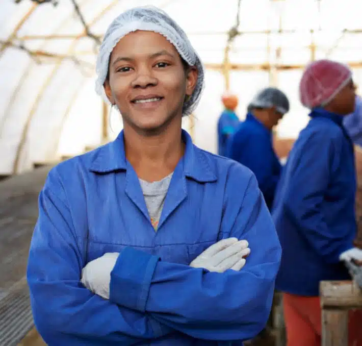A factory worker in a blue uniform and hairnet smiles with arms crossed, embodying the efficiency of cloud-basiertes PLM für die Lebensmittel- und Getränkeindustrie. In the blurred background, colleagues in blue uniforms bustle about within the expansive workspace.