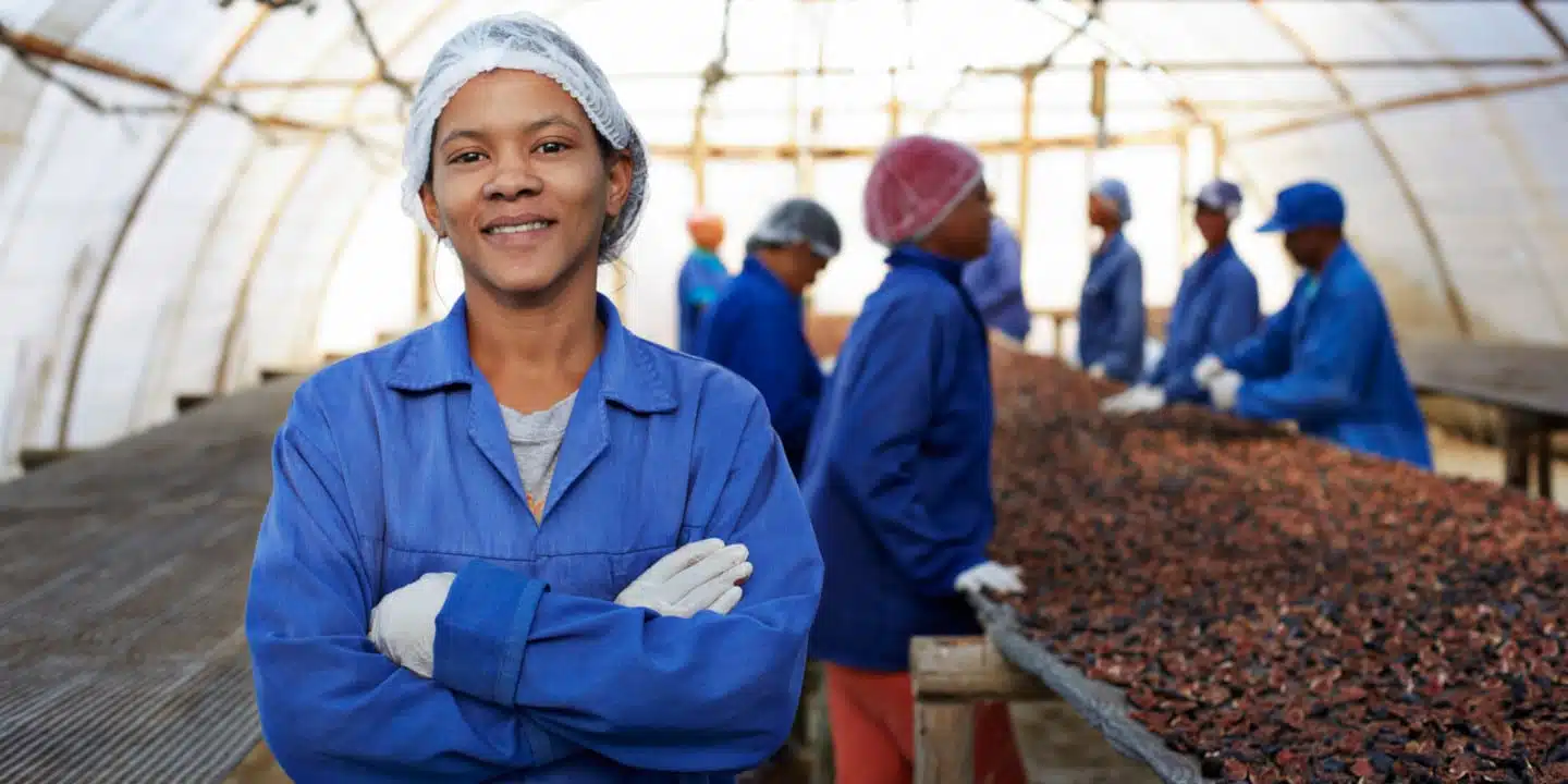 A person in a blue uniform and hair net smiles with arms crossed in a greenhouse, overseeing a cloud-based PLM system while others work with dried crops on tables in the background.